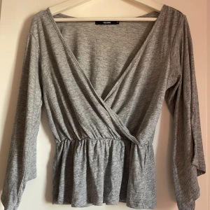 Grå omlottopp från Bik Bok - Grå långärmad omlottopp från Bik Bok med v-ringning och resår i midjan. Tunt och mjukt material.