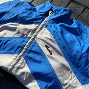 Vindjacka från Ellesse - Säljer en retro vindjacka från Ellesse i blått, grått och vitt med dragkedja och hög krage. Jackan har ett sportigt mönster och Ellesse-logga på bröstet. Perfekt för dig som gillar vintage streetwear. Storleken är 50 vilken är ungefär L