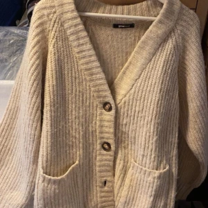 Beige stickad kofta från Gina Tricot - Mysig beige stickad kofta från Gina Tricot med stora knappar framtill och två fickor. Koftan har lång ärm och bred ribbad kant vid halsen och längs knäppningen. Perfekt för lager-på-lager under kyliga dagar.