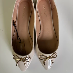 Beige ballerinaskor med guldfärgad rosett från Anna Field - Beige ballerinaskor från Anna Field med spetsig tå och dekorativ guldfärgad rosett framtill. Säljer pga förstora och de är i storlek 37 men passar för 38/39. Aldrig använda