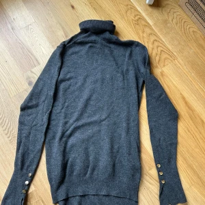 Mörkgrå polotröja från Zara - Säljer en mörkgrå polotröja från Zara med långa ärmar och snygga guldfärgade knappar vid ärmsluten. Tröjan är stickad och har en klassisk, stilren look som passar perfekt till många outfits.