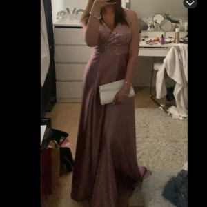 Lång glittrig/satin rosa balklänning - Säljer en elegant långklänning i rosa med glittrig överdel och v-ringning. Klänningen har axelband och ett slätt, flytande kjolparti i satinliknande material. Perfekt för bal eller festliga tillfällen. Den är endast använd en gång! Pris kan diskuteras vid snabb affär!! 