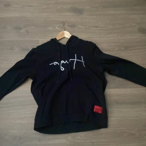 Svart hoodie från Hugo - Säljer en svart hoodie från Hugo med vit logga framtill och röd Hugo-lapp nedtill. Tröjan har huva med snörning och långa ärmar. Perfekt för en avslappnad och stilren look. Storlek M. 