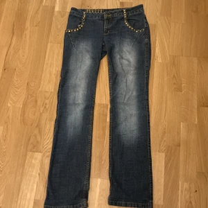 Blå low waisted jeans med nitar från Brooker - Snygga blå low waisted jeans från Brooker med raka ben och coola guldfärgade nitar längs fickor och linning. Klassisk femficksmodell med slitningar framtill och bak. Perfekta för dig som vill sticka ut lite extra.