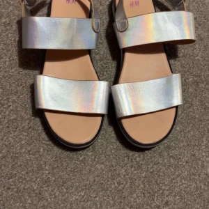 Silvriga sandaler från H&M - Säljer ett par trendiga sandaler från H&M i metallic-silver med breda remmar och kardborreknäppning. Skorna har en platt, svart räfflad sula och öppen tå. Perfekta för att ge outfiten en futuristisk touch!