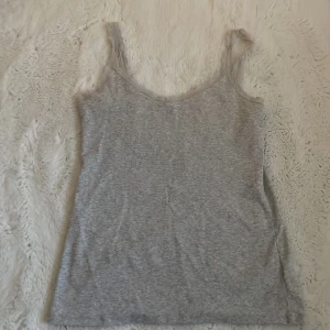Grå ribbad linnetopp med spets från H&M - Säljer en ljusgrå ribbad linnetopp från H&M med tunna axelband och fina spetsdetaljer längs kanterna. Perfekt att styla till jeans eller kjol för en enkel och snygg look.