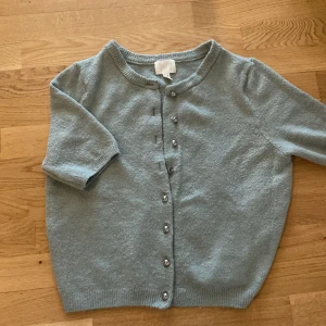 Ljusblå kofta från H&M - Säljer en ljusgrön kofta från H&M i storlek M. Koftan har korta ärmar och silverfärgade knappar framtill. Materialet känns mjukt och passar perfekt för lager-på-lager. Enkel och stilren design som funkar till mycket.