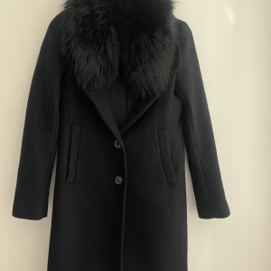 Svart kappa med fuskpäls från Zara Woman - Säljer en elegant svart kappa med avtagbar päls från Zara  med stor, fluffig fuskpäls runt kragen. Kappan har två knappar framtill och raka fickor. Perfekt för dig som vill ha en stilren och klassisk look.