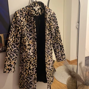 Leopardmönstrad kappa i gult och svart - Säljer en snygg kappa med leopardmönster i svart, gult och vitt. Jackan har lång ärm, krage och dolda fickor på sidorna. Perfekt statement-plagg för dig som vill sticka ut med din stil. Slitstarkt tyg 