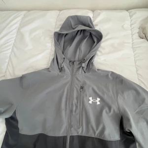 Grå vindjacka med huva från Under Armour - Snygg grå vindjacka från Under Armour med huva och dragkedja. Jackan har en vit logga på bröstet och en bröstficka med dragkedja. Perfekt för utomhusaktiviteter och träning.