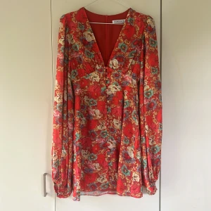 Röd blommig klänning från Adoore - Säljer en röd blommig klänning från Adoore med v-ringning och långa ärmar. Klänningen har ett färgglatt blommönster i beige, blått och grönt samt markerad midja och lätt utsvängd kjol. Perfekt för vår och sommar! Säljer då den tyvärr blivit förliten andvänd endast ett fåtal gånger 