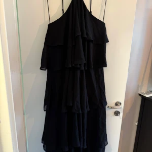 Svart långklänning med volanger - Säljer en svart långklänning med flera lager volanger och asymmetrisk one shoulder-skärning. Klänningen har en elegant och flytande siluett, perfekt för speciella tillfällen. Snygg och stilren design som sticker ut.