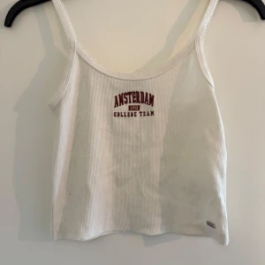 Vit ribbad linnetopp med tryck - Säljer en vit ribbad linnetopp med texten 'AMSTERDAM COLLEGE TEAM' i rött framtill. Tunt material och smala axelband.
