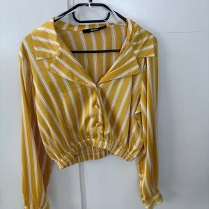 Gul- och vitrandig skjorta från BikBok - Säljer en gul- och vitrandig skjorta från BikBok i storlek XS. Skjortan har långa ärmar, knappar framtill och en croppad passform med resår i midjan. Perfekt för dig som gillar färg och mönster!