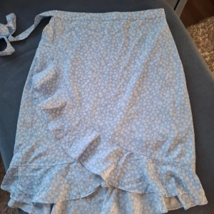 Ljusblå blommig omlottkjol från Vero Moda - Supersöt ljusblå omlottkjol från Vero Moda med vitt blommönster och volangdetaljer längs kanten. Kjolen knyts i midjan!