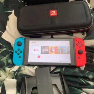 Nintendo Switch med fodral och handkontroll - Säljer en Nintendo Switch med blå och röd Joy-Con, svart bärväska med Nintendo Switch-logga och en svart handkontrollhållare. Perfekt för gaming hemma eller på resan.  fodral ingår.