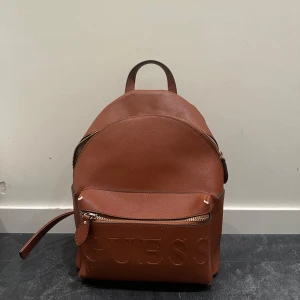 Brun ryggsäck från Guess - Säljer en snygg brun ryggsäck från Guess med präglad logga framtill och guldfärgad dragkedja. Väskan har ett stort huvudfack och en ytterficka med dragkedja. Insidan är fodrad med mönstrat tyg och har flera fack för småsaker. 