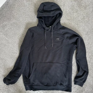 Svart hoodie från Under Armour, herr M - Säljer en svart hoodie från Under Armour med klassisk logga på bröstet. Tröjan har huva med dragsko och en stor magficka framtill. Lite urtvättad men egentligen inget som stör, ser mer gråsvart ut bara. Herr stl M.
