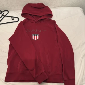 Röd hoodie från GANT - Säljer en röd hoodie från GANT med broderad logga och sköld på bröstet. Tröjan har huva och en stor magficka framtill. Perfekt för dig som gillar klassisk och sportig stil.