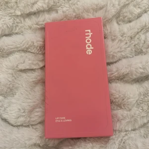 Rhode Lip Case i rosa IPHONE 14 pro - Säljer ett rosa Rhode Lip Case, ett mobilskal med plats för läpprodukt. Skalet är gjort i mjukt silikonmaterial och har en unik design med Rhode-logga. Perfekt för att alltid ha läpprodukten nära till hands.