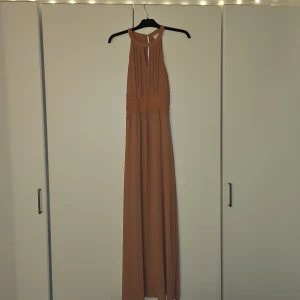 Beige / gammelrosa långklänning med spetsdetaljer - Säljer en elegant beige långklänning med vackra spetsdetaljer i midjan och hög halsringning. Klänningen är ärmlös och har en lätt, flowy passform som ger en stilren och sofistikerad look.