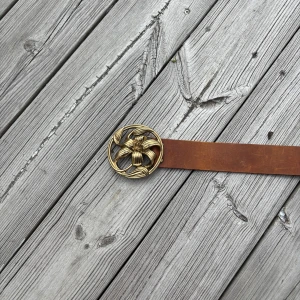 Brunt bälte med stor guldfärgad blomspänne - Säljer ett brunt bälte med ett stort, runt spänne i guldfärg med blommönster. Bältet är i skinn och har en unik och iögonfallande design som passar perfekt för att lyfta en outfit.