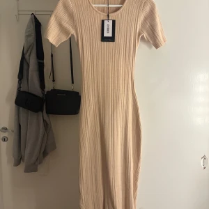 Beige ribbad klänning från 4th & Reckless - Beige ribbad klänning från 4th & Reckless med kort ärm, rund halsringning och öppen rygg. Klänningen har knappar längs sidan och är figurnära med en snygg slitsdetalj. Aldrig använd och prislapp finns kvar. 