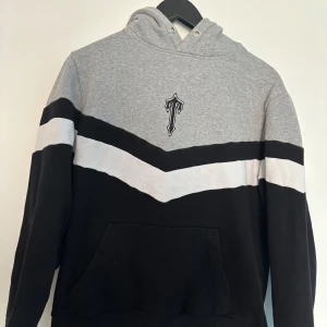 Grå och svart hoodie från Trapstar - Säljer en snygg hoodie från Trapstar i grått och svart med vit och svart diagonal rand över bröstet. Tröjan har broderad logga på bröstet. 