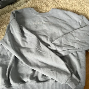 Ljusgrå sweatshirt med text - Säljer en ljusgrå sweatshirt med rund halsringning och trycket 'CENTRE OF ATTENTION' framtill. Tröjan har långa ärmar och ribbade muddar vid ärmslut och nederkant. Perfekt för en avslappnad stil.