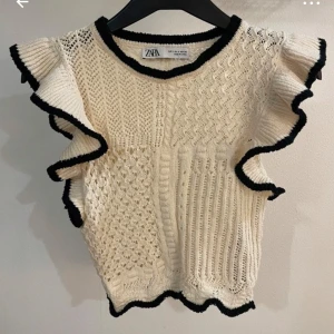Stickad topp med volang från Zara - Säljer en krämvit stickad topp från Zara med svarta kanter och volangdetaljer längs ärmhålen. Toppen har ett vackert stickat mönster och rund halsringning. Perfekt för att lyfta en outfit!