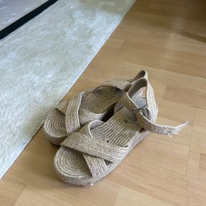 Sandlatter med klack - Säljer ett par beige sandaletter med platåsula som utgör en klack. Skorna har en öppen tå och justerbart spänne vid ankeln. Materialet är flätad textil som ger en naturlig och somrig känsla. Mycket bekväma att gå i. Märkta med storlek 37 men utmärkt för 38.