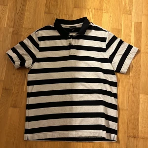 Svartvit randig pikétröja från H&M - Säljer en klassisk svartvit randig pikétröja från H&M. Tröjan har korta ärmar, krage och knäppning framtill. Perfekt för en avslappnad och stilren look. Passar dig som gillar enkel och tidlös stil.