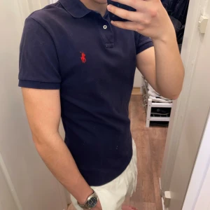 Polo Ralph Lauren (Blå piké) - Snygg blå piké från Polo Ralph Lauren med klassisk krage och knappar framtill. Ikonisk röd broderad logga på bröstet. Perfekt till jeans eller shorts för en stilren look. Den är oanvänd och redo för en ny ägare! Pikén är storlek S!