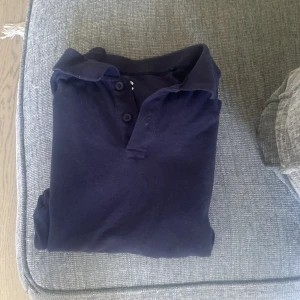 Mörkblå långärmad pikétröja - Säljer en klassisk mörkblå långärmad pikétröja med krage och knappar vid halsen. Perfekt för en stilren och avslappnad look. Passar både till jeans och chinos.