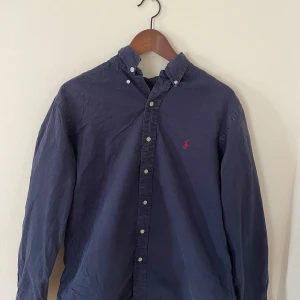 Mörkblå skjorta från Ralph Lauren - Klassisk mörkblå skjorta från Ralph Lauren röd broderad logga på bröstet. Storlek L men passar som M | Skick 9/10.  Skjortan har lång ärm och knäppning framtill. Perfekt till en stilren och avslappnad look. 