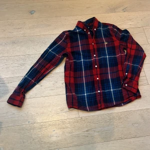 Rutig skjorta från Gant - Snygg röd och blå rutig skjorta från Gant med klassisk krage och vita knappar. Skjortan har lång ärm och broderad Gant-logga på bröstet. Perfekt för dig som gillar en tidlös och stilren look.