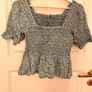 Blommig blus med puffärm - Supersöt blommig blus i grön och vitt med smockad överdel och volang nertill. Puffiga korta ärmar och liten rosett framtill. Perfekt till jeans eller kjol för en trendig look.