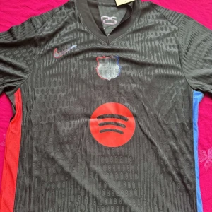 FC Barcelona svart fotbollströja Nike - Säljer en svart FC Barcelona fotbollströja från Nike med klubbmärke och röd Spotify-logga på bröstet. Tröjan har röda och blå detaljer på sidorna och Lamine Yamal 19 tryckt på ryggen. Perfekt för dig som älskar Barca!