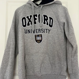 Grå Oxford University hoodie - Grå hoodie med svart och vit Oxford University-tryck framtill samt broderad logga. Stor ficka på magen och huva med dragsko. Liten brittisk flagga på ärmen. Perfekt för en avslappnad stil.
