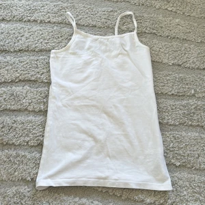 Vit enkelt linne med smala axelband - Säljer en vit, linne med smala axelband och rak passform. Perfekt för varma dagar eller som basplagg. Mjuk och lätt i materialet, enkel att styla med olika accessoarer.