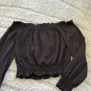 Svart offshoulder blus - Säljer en svart offshoulder blus med långa ärmar och smockdetaljer vid hals, midja och ärmslut. Blusen har en lätt volangkant upptill och nertill för en extra snygg touch.