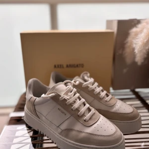 Vita sneakers från Axel Arigato - Säljer ett par stilrena vita sneakers från Axel Arigato med beige detaljer. Skorna har snörning, perforerad tå och en platt sula. Perfekta för dig som gillar minimalistisk design och hög komfort.