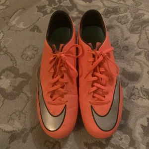 Nike Mercurial fotbollsskor  - Säljer ett par orangea Nike Mercurial fotbollsskor med svart och silverfärgad swoosh. Skorna har snörning och dobbar på sulan för bra grepp på planen. Perfekta för dig som vill sticka ut på fotbollsplanen!