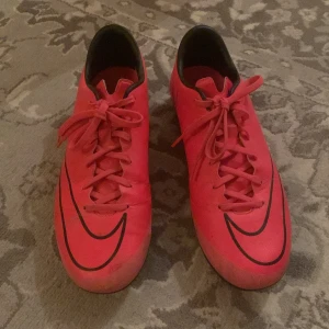 Nike Mercurial fotbollsskor  - Säljer ett par Nike Mercurial fotbollsskor i neonrosa med svarta detaljer och snörning. Skorna har rund tå, platt sula med dobbar och är designade för spel på gräs. Perfekta för dig som vill sticka ut på planen!
