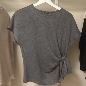 Grå topp med knytdetalj från Zara - Säljer en stilren grå topp från Zara med korta ärmar och snygg knytdetalj i sidan. Toppen har rund halsringning och är tillverkad i ett mjukt material som känns bekvämt mot huden. Perfekt för en enkel men trendig look.