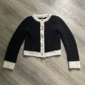 Svart fluffig kofta med vita detaljer - Säljer en svart fluffig kofta med vita kanter och glittriga knappar. Jätte skönt och gosig tyg. Säljer den för den inte rigitt är min stil