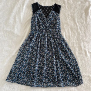 Blommig omlottklänning från Vero Moda - Supersöt blommig klänning från Vero Moda i storlek XS. Klänningen har omlottliv, spetsdetaljer vid axlarna och är ärmlös. Mönstret är småblommigt i blått, vitt och svart. Perfekt för vår och sommar! Har några lösa trådar. Längd: 88cm