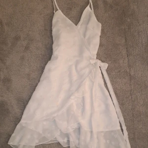 Vit omlottklänning med volang och knytband - Säljer en söt vit omlottklänning med smala axelband och volangdetaljer. Klänningen har knytband i midjan och är dekorerad med diskreta broderade blommor. Perfekt för sommarens alla tillfällen!