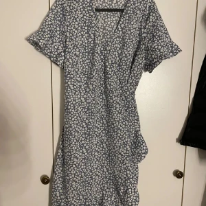 Blommig omlottklänning från Vero Moda - Söt blå och vit blommig omlottklänning från Vero Moda med korta ärmar och volangdetalj nertill. Klänningen har knytband i midjan och är perfekt för vår och sommar. Lätt och luftig modell som ger en avslappnad känsla.