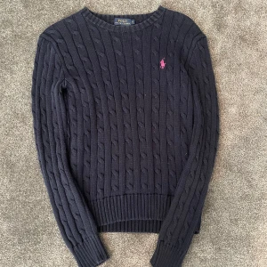 Mörkblå stickad tröja från Polo Ralph Lauren - Mörkblå kabelstickad tröja från Polo Ralph Lauren med rund halsringning och rosa broderad logga på bröstet. Klassisk och tidlös modell med ribbade muddar vid ärmslut och nederkant. Perfekt för lager-på-lager-stil.